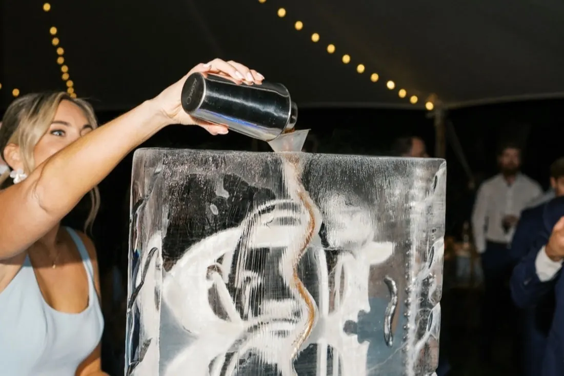 A guest pouring from a shaker through a carved ice luge under a marquee strung with festoon lights