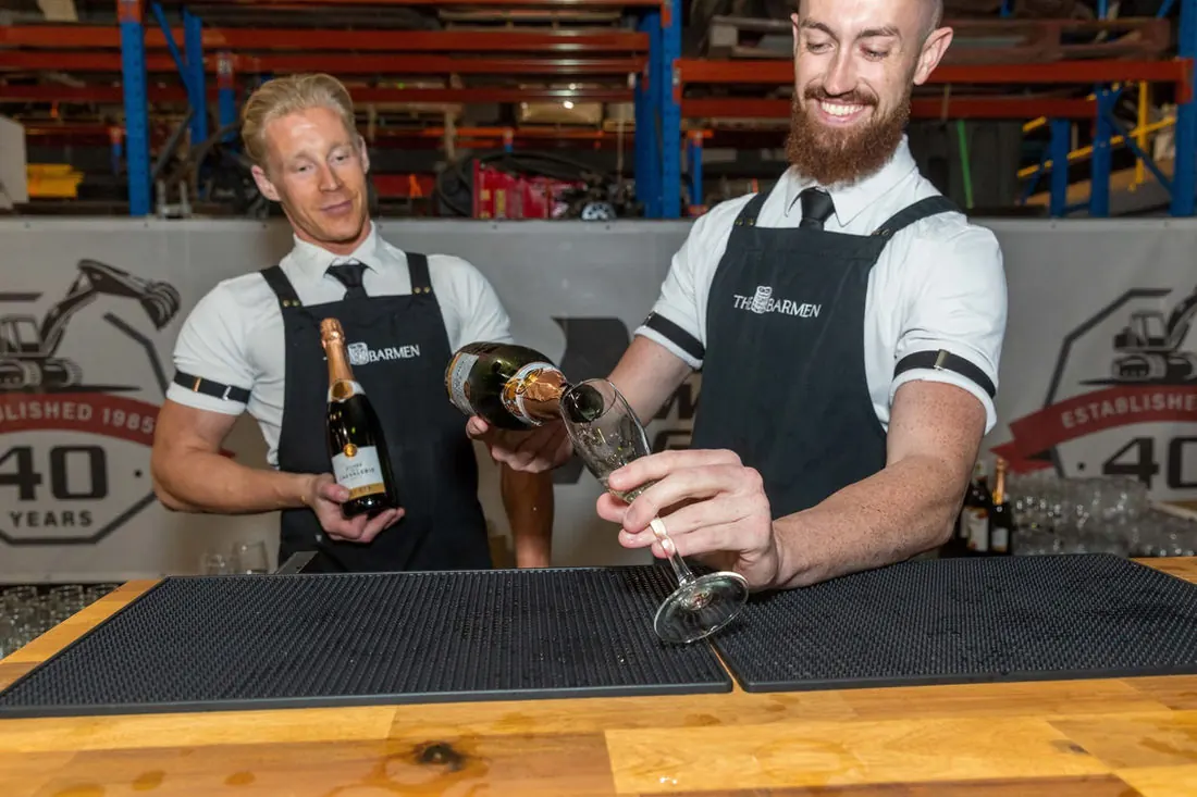 Two Barmen bartenders in branded aprons behind a timber bar, one pouring sparkling wine into a flute and the other holding the bottle