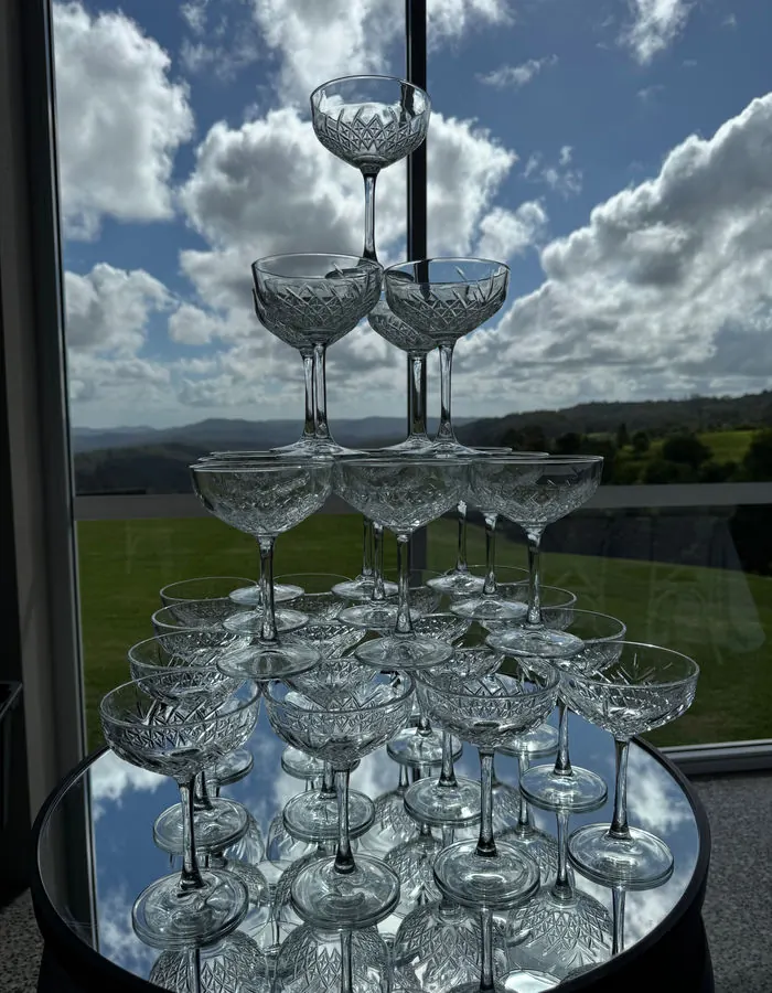 A tower of cut-crystal champagne coupes stacked on a mirrored table in front of a window with hinterland views