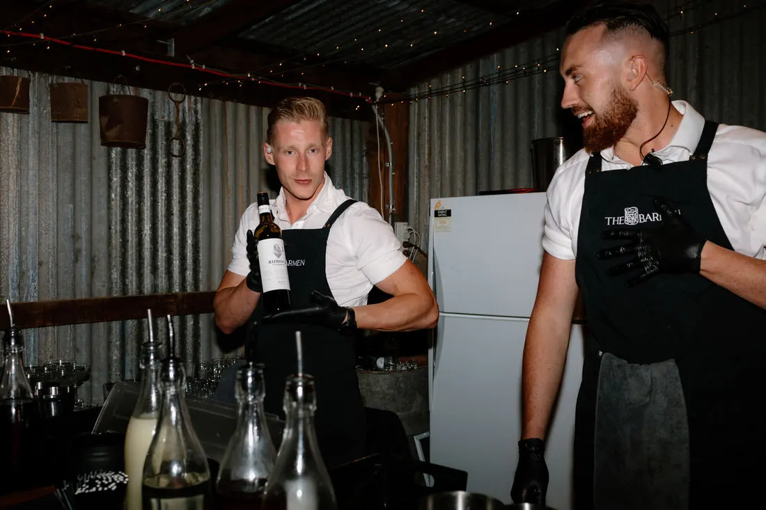 Two Barmen bartenders in branded aprons working side by side at a rustic bar lined with bottles