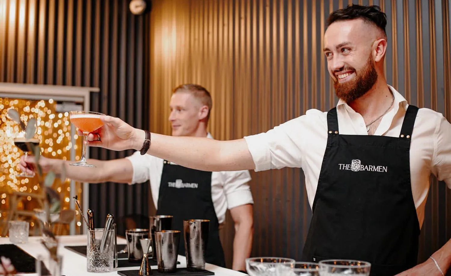 A Barmen bartender in a white shirt and black apron handing a finished cocktail across a warm, softly lit bar while a colleague works behind him