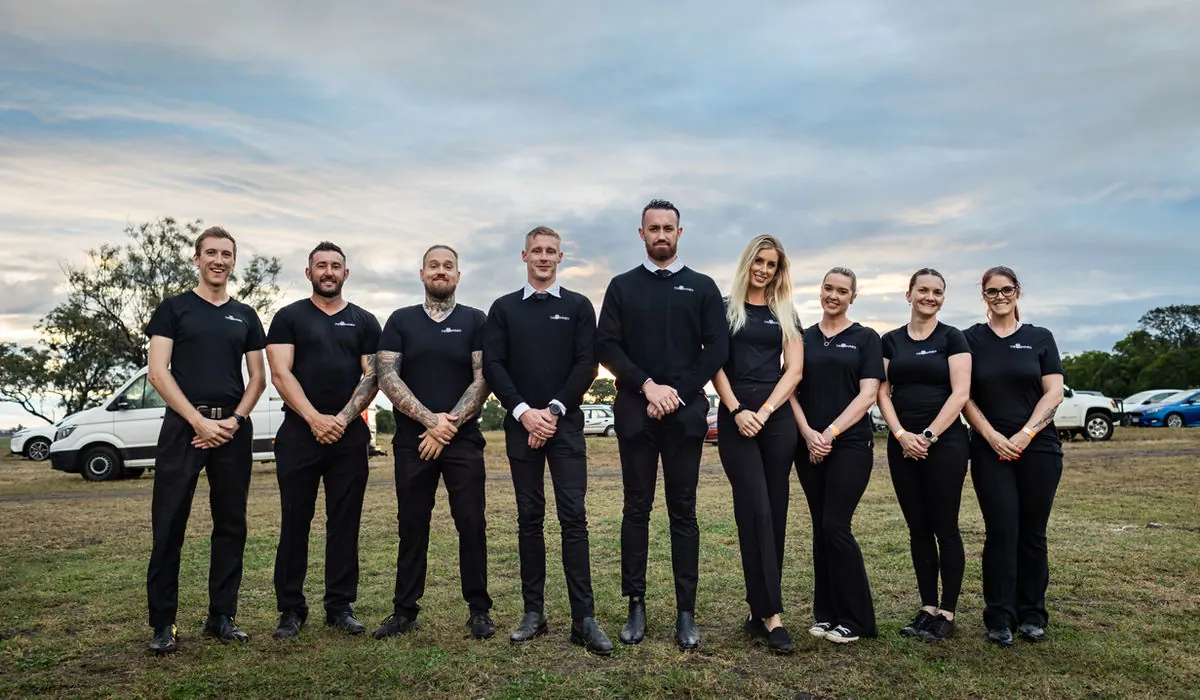 The Barmen team of ten in matching black uniform standing in a line on grass before an event, with trees and sky behind