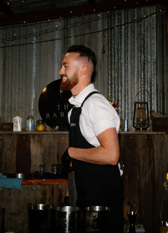 A laughing Barmen bartender in a white shirt and black apron leading a session at a rustic timber bar beneath The Barmen sign