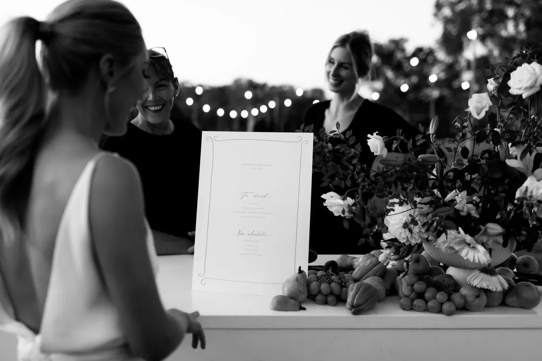 Black and white photograph of a bride in white beside a grazing table dressed with a printed menu sign, guests chatting under festoon lighting
