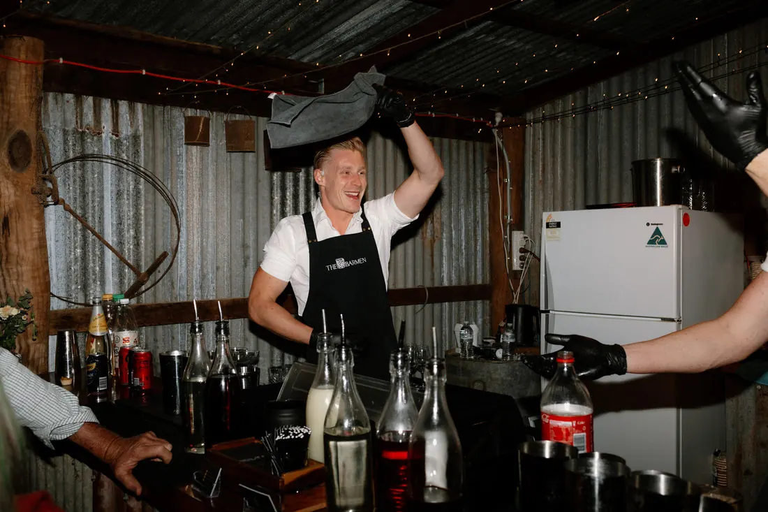 A Barmen bartender lifting an esky overhead behind a bar in a corrugated-iron shed, bottles of spirits and syrups lined up in front of him