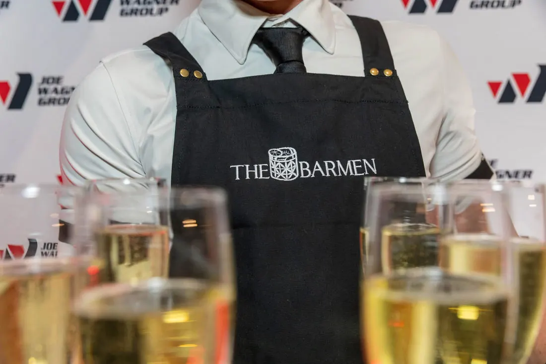 Close-up of a Barmen bartender's black apron embroidered with The Barmen logo behind a row of freshly poured champagne flutes at a corporate media wall