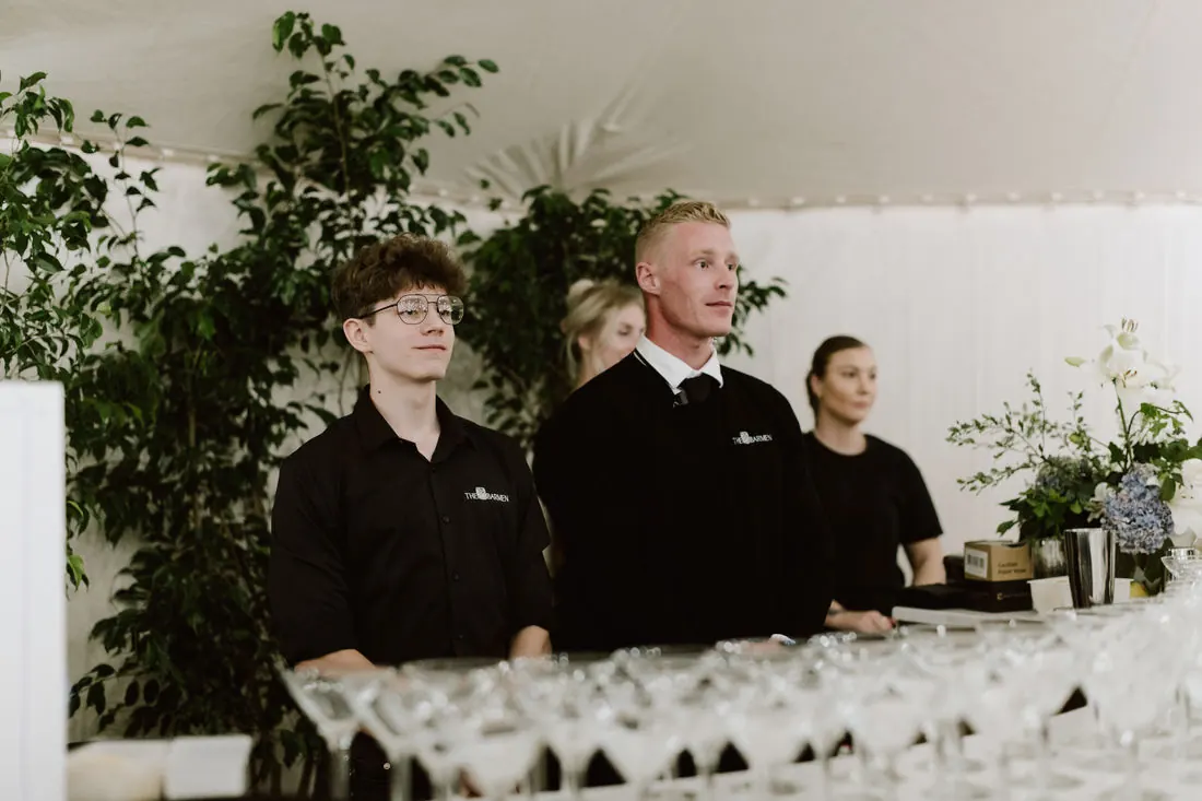 Four Barmen bartenders in black uniform standing behind a bar lined with rows of champagne coupes inside a white marquee, greenery beside them