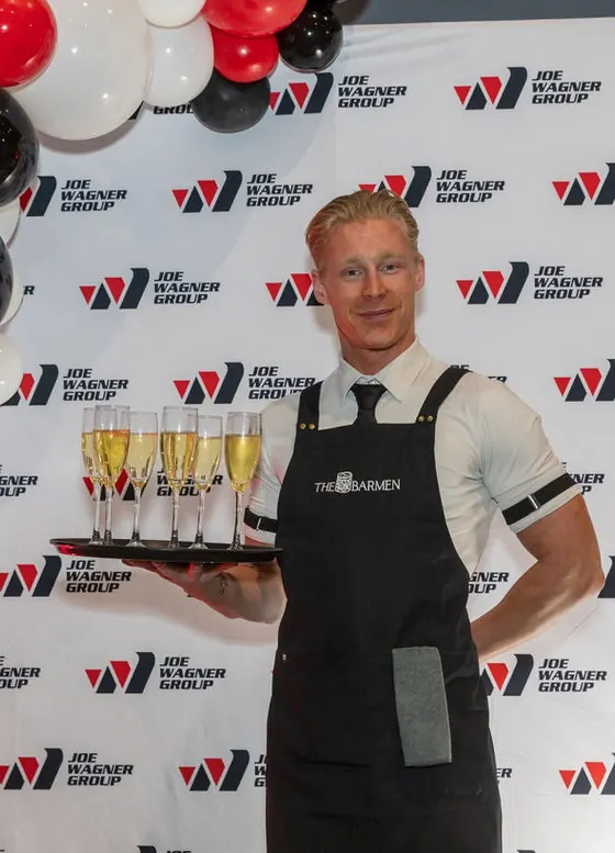 A Barmen bartender in a black apron holding a tray of champagne flutes in front of a branded corporate media wall with balloons