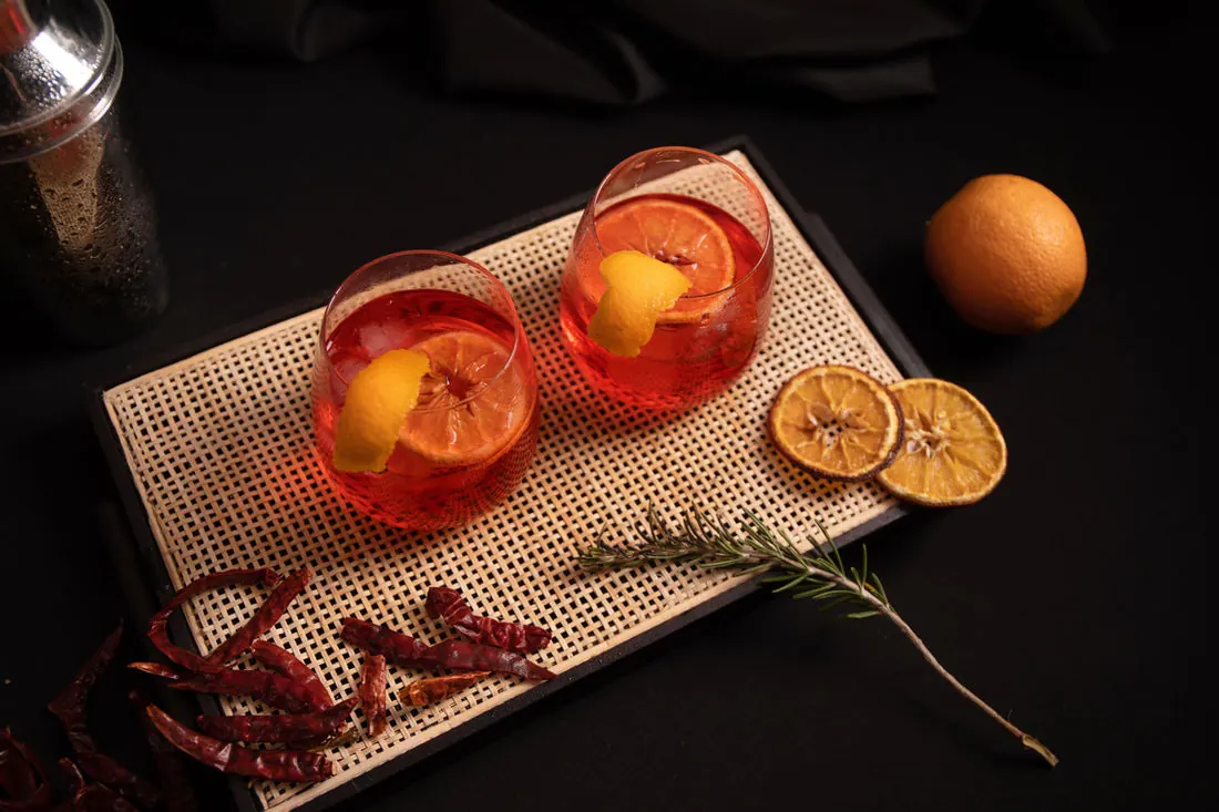 Two spritz cocktails on a rattan tray finished with dehydrated orange wheels, a fresh orange and rosemary beside them
