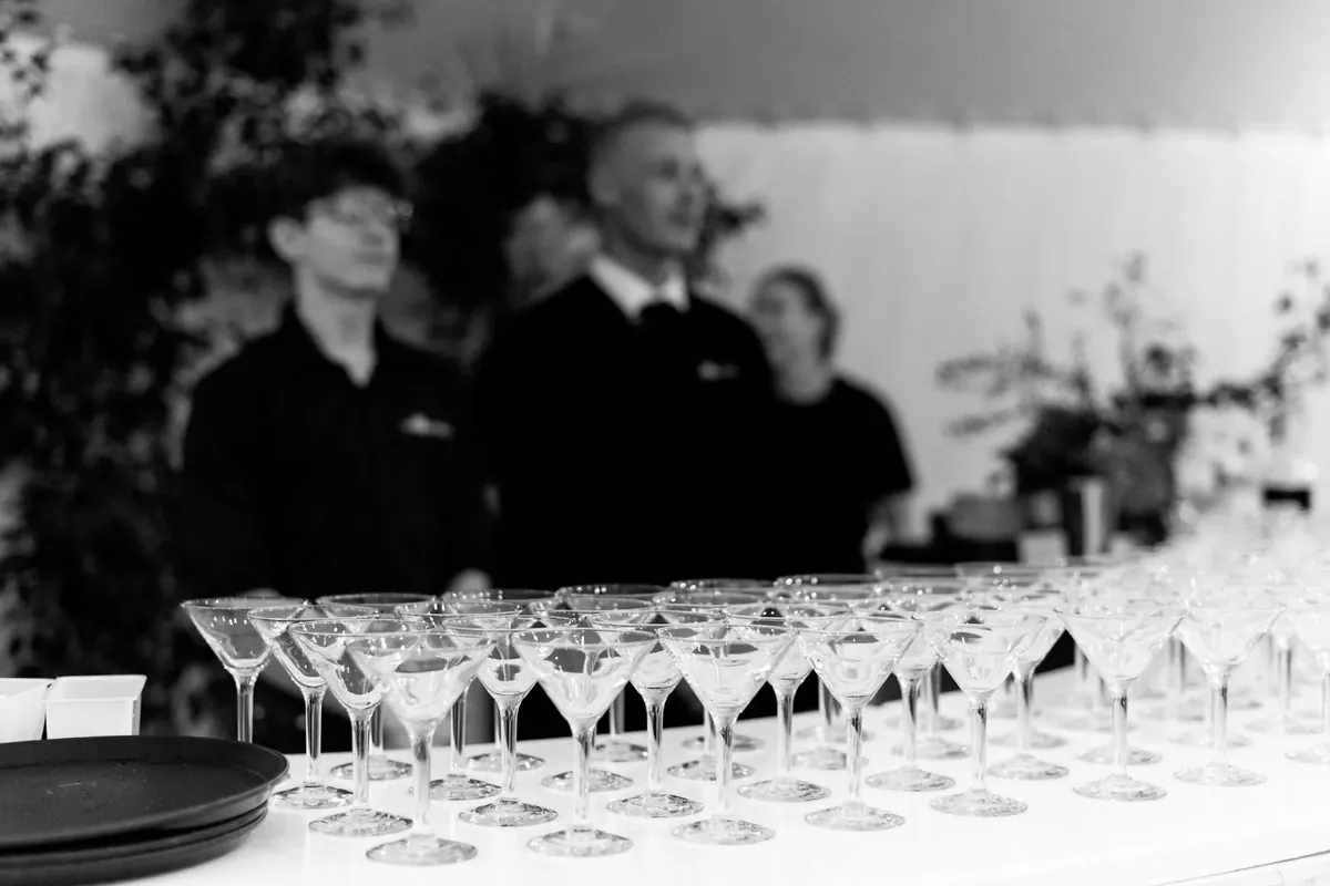 Black and white photograph of the Barmen team in matching black uniform standing behind long rows of polished champagne coupes before an event