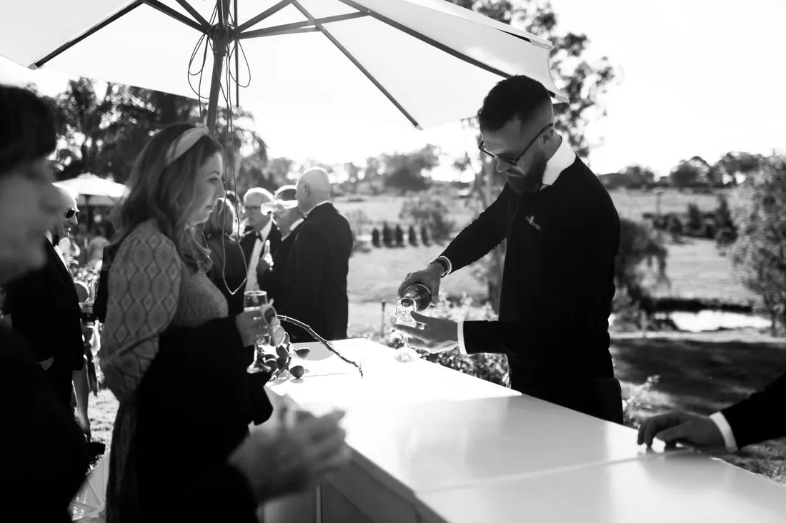 Black and white photograph of a Barmen bartender serving guests at a white outdoor bar under a large umbrella at a daytime event