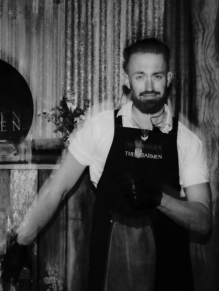 Black and white portrait of a Barmen bartender in a branded apron and gloves at a timber bar beneath a round The Barmen sign