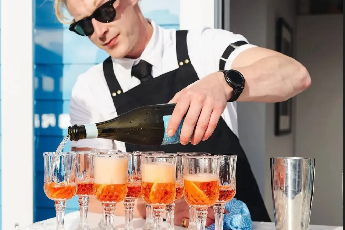 A Barmen bartender in sunglasses pouring sparkling wine into a row of coupes filled with orange cocktails outside a corporate venue